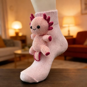 May include: A light pink sock with a crocheted axolotl toy attached. The axolotl is pink with dark pink accents and black eyes. The sock is made of a soft, knitted material. The axolotl toy is approximately 3 inches tall.