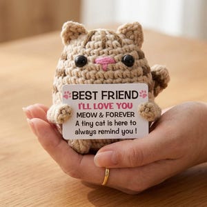 May include: A crocheted cat figurine in shades of tan and brown, holding a white card. The card reads "BEST FRIEND, I'LL LOVE YOU, MEOW & FOREVER, A tiny cat is here to always remind you!" The cat has black eyes and a pink nose.