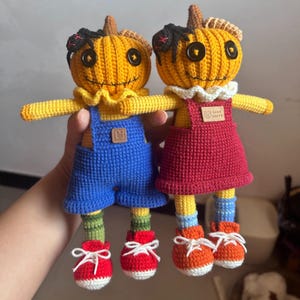 May include: Two handmade crochet dolls with pumpkin-shaped heads, button eyes, and stitched smiles. One doll wears blue overalls, the other a red dress. Both have yellow arms, striped socks, and red or orange shoes. The dolls are holding hands.