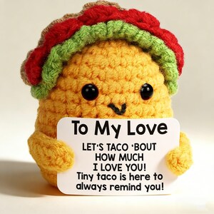 May include: A crocheted taco plushie with a smiling face and a red, green, and brown shell. The taco holds a white sign that reads "To My Love, Let's Taco 'Bout How Much I Love You! Tiny taco is here to always remind you!"