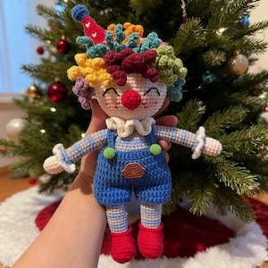 May include: A handmade crochet clown doll with a colorful wig, red nose, and smiling face. The clown wears blue overalls with green buttons and red shoes. The doll is approximately 8 inches tall and is set against a Christmas tree background.