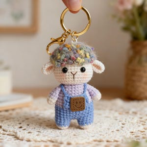May include: A handcrafted crochet sheep keyring. The sheep features a white face, ears, and body, topped with a colourful hat. It's dressed in blue dungarees with a smiling face patch. The keyring has a gold-coloured ring and clasp.