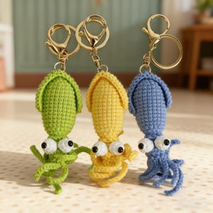 May include: Three colourful crochet squid keyrings in green, yellow, and blue. Each squid has large googly eyes and gold-coloured metal key rings. The tentacles are curled, and the keyrings are displayed against a soft background.