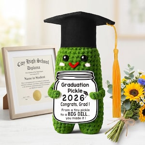 Puede incluir: Un pepinillo verde tejido a ganchillo con un gorro de graduación negro y una borla amarilla. El pepinillo sostiene un cartel que dice "Graduation Pickle 2026 Congrats, Grad! From a tiny pickle to a BIG DILL... you made it!" Un diploma y flores en el fondo.