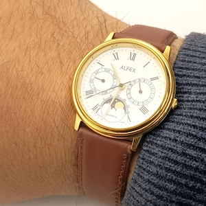 Peut inclure: Une montre ALFEX à quartz dorée avec un cadran blanc, des chiffres romains et un bracelet en cuir marron. Le cadran affiche les indicateurs de jour, de date et de phase lunaire. Le bracelet est en cuir marron.