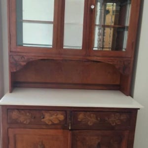 Può includere: Un mobile in legno con ante a vetri e piano bianco. Il mobile ha due cassetti e due ante con disegni intagliati a foglie e fiori. La sezione superiore ha ante a vetri e uno schienale a specchio.