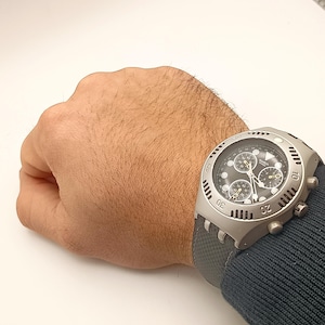 May include: A Swatch chronograph watch with a gray dial and a black strap. The watch has a stainless steel case and features multiple subdials. The watch is on a wrist with a dark blue sleeve.