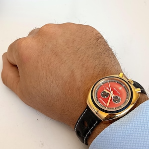 Peut inclure: Une montre dorée avec un cadran rouge et des sous-cadrans noirs. La montre a un bracelet en cuir noir avec des coutures blanches. Le cadran affiche le texte "JACKY ICKX" et "EASY RIDER".
