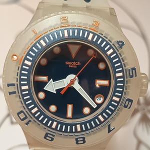 May include: A transparent Swatch watch with a navy blue dial and orange accents. The watch features a white arrow-shaped hand and a date display at the 3 o'clock position. The watch band is transparent.
