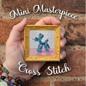 May include: A small, gold-framed cross-stitch featuring a blue balloon dog on a pink and white background. The frame is held in a hand against a brick wall. Text reads "Mini Masterpiece" and "Cross Stitch".