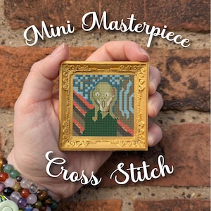 Pode incluir: Uma pequena obra de arte em ponto cruz de uma pintura famosa, emoldurada em uma moldura dourada. A obra de arte apresenta uma figura com a boca aberta e os olhos arregalados. O texto "Mini Masterpiece Cross Stitch" é exibido acima e abaixo.