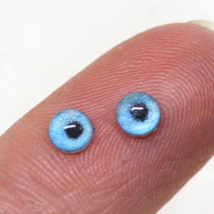 May include: Two small blue plastic doll eyes with black pupils. The eyes are on a finger.