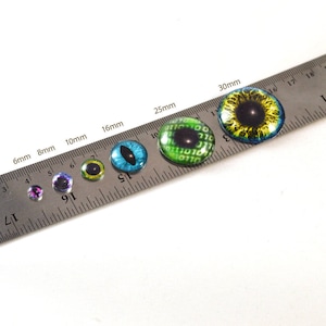 May include: A ruler with a set of colorful plastic eyes in different sizes, ranging from 6mm to 30mm. The eyes have various designs, including a blue cat eye, a green eye with a binary code pattern, and a yellow eye with a black pupil.
