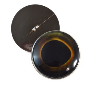 May include: A round, black button with a silver rim and a realistic eye design. The eye has a black pupil, a yellow iris, and a brown outer ring.