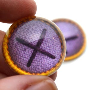 May include: Two round, glass cabochons with a purple fabric pattern and a black cross design. The edges have a gold and orange border. One cabochon is in focus, held by a person's fingers, with another slightly blurred in the background.