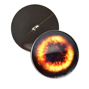 May include: Two round buttons. One button features a detailed eye design with fiery orange, yellow, and black hues. The other button is a solid dark brown color with a metal loop on the back for sewing. The buttons are on a white background.
