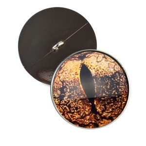 May include: A round button with a detailed image of a golden eye with a black pupil. The back of the button is black.