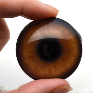 May include: A close-up of a brown glass eye with a black pupil. The eye is being held by a finger.