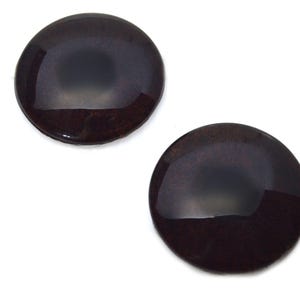Op de afbeelding: Twee donkerbruine, glanzende, ronde cabochons met een reflecterend oppervlak. De cabochons hebben een licht gewelfde vorm en zijn geplaatst tegen een witte achtergrond. Dit zijn waarschijnlijk knutselbenodigdheden voor het maken van sieraden of andere creatieve projecten.