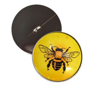 May include: A round button with a yellow background and a detailed illustration of a bee. The bee is black and orange with detailed wings. The button has a silver metal rim and a dark brown back with a metal loop.