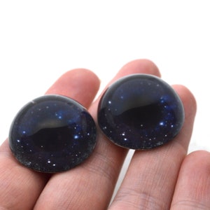May include: Two round black and blue glass eyes with a galaxy print. The eyes have a dark blue centre with white stars scattered across the surface.