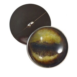 以下が含まれることがあります： 銀色の金属製の縁取りと黄色、茶色、黒の目のデザインが施された丸いボタン。ボタンの裏側は黒です。