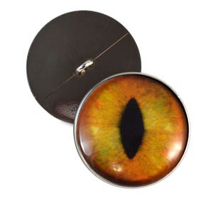 May include: A round button with a yellow and orange cat eye design. The button has a silver rim and a black pupil.