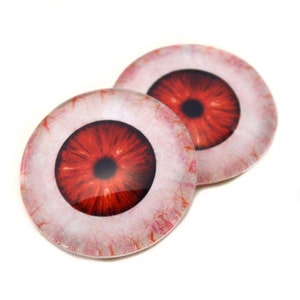 May include: Two round, glass eye cabochons with a realistic, bloodshot design. The irises are a deep red with black pupils, surrounded by a white sclera detailed with red veins. These craft supplies are ideal for creating unique jewelry or art projects.