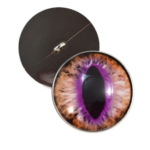 May include: A round, purple and orange eye button with a black pupil. The button is surrounded by a silver metal rim.