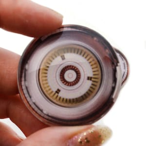 May include: A round, glass, decorative paperweight with a detailed image of a circular, metallic, industrial design. The design features concentric circles and a central hub with a red ring.