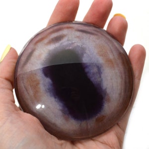 May include: A round, polished agate stone with a purple and brown swirl pattern. The stone is translucent and has a glossy finish.