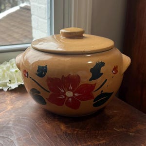May include: Tan ceramic lidded pot with a floral design. The pot features a large red flower with black and green accents. The pot has two handles and sits on a wooden surface. The lid has a small knob.