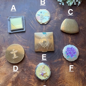 May include: A collection of vintage compact mirrors in various shapes and designs. The mirrors are gold, silver, and have decorative elements. One mirror is square, another round with a floral design, and others have intricate patterns. The collection is displayed on a wooden surface.