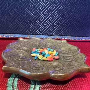 Puede incluir: Un plato decorativo de metal de estilo antiguo con un diseño floral ornamentado. El plato contiene una colección de semillas coloridas pintadas en tonos azules, amarillos, naranjas y rojos. El plato está sobre una superficie roja.