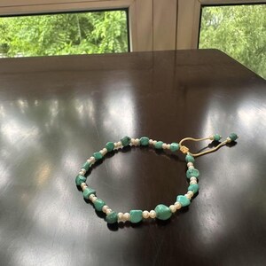 May include: A turquoise and white beaded bracelet. The bracelet is made with irregular turquoise stones and small white beads. It has a beige adjustable cord closure. The jewellery is displayed on a dark, reflective surface.