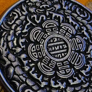 May include: A detailed, silver-colored pendant with an intricate design. The pendant features a central circular emblem surrounded by ornate patterns and stylized figures, possibly dragons. The design is raised, creating a textured surface.