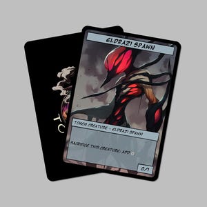 May include: Two fantasy trading card game cards. One card features a creature with a red head and black tendrils, labeled "ELDRAZI SPAWN." The other card is black with a white graphic. The cards are on a gray background.