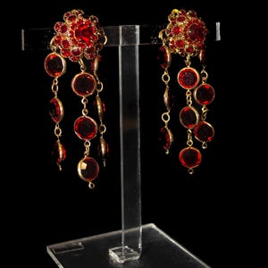 Op de afbeelding: Een paar sierlijke rood-gouden oorbellen. Elke oorbel heeft een bloemvormige cluster van rode edelstenen aan de bovenkant, met meerdere hangende rode cirkelvormige edelstenen. De oorbellen worden getoond op een heldere acryl standaard tegen een zwarte achtergrond.