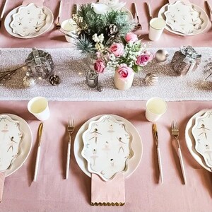 May include: A festive table setting with ballet-themed plates, gold cutlery, and pink napkins. The centerpiece features a floral arrangement and decorative ornaments. The table is set on a pink tablecloth, creating an elegant and celebratory atmosphere.