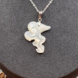 Puede incluir: Un collar de plata con un colgante que presenta un &aacute;ngel tallado en perla suave. El &aacute;ngel tiene alas detalladas y una expresi&oacute;n serena. El texto "SOFT PEARL" se muestra en la parte inferior de la imagen.