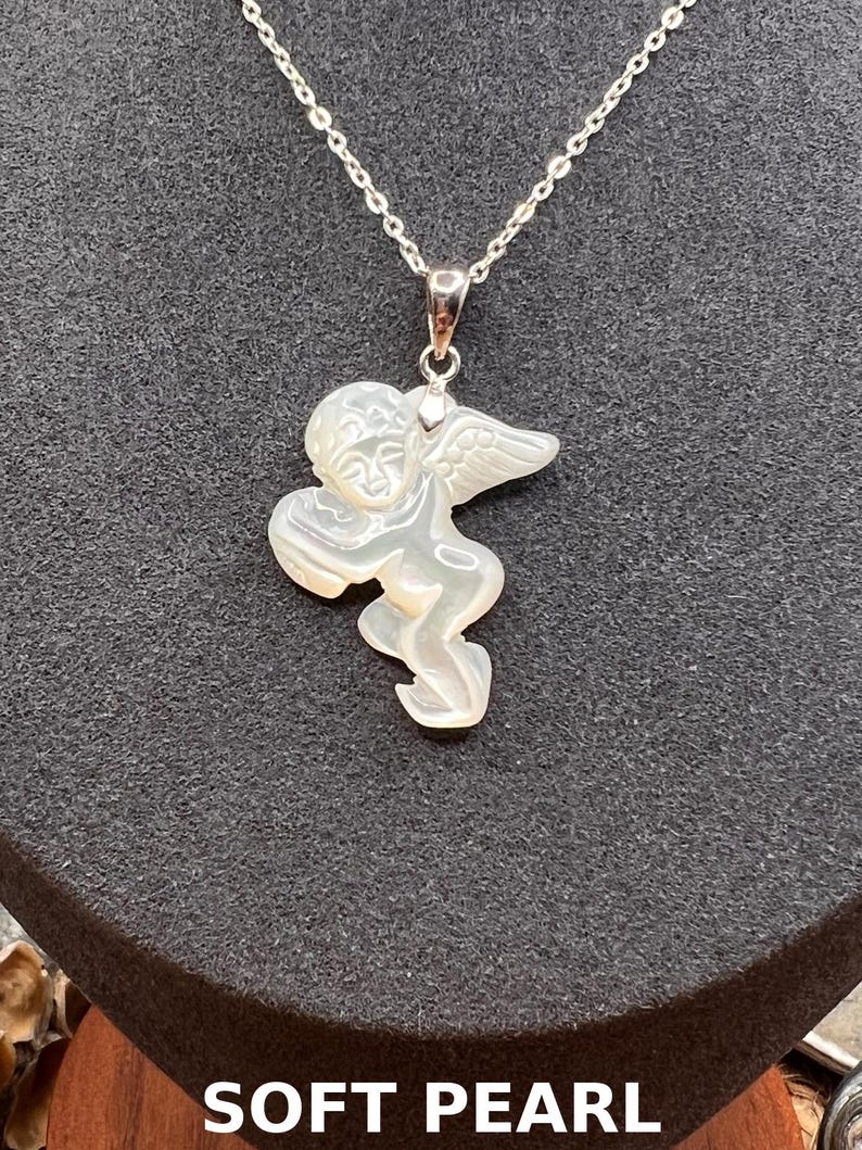 Puede incluir: Un collar de plata con un colgante que presenta un &aacute;ngel tallado en perla suave. El &aacute;ngel tiene alas detalladas y una expresi&oacute;n serena. El texto "SOFT PEARL" se muestra en la parte inferior de la imagen.