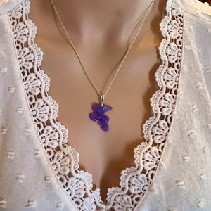 Puede incluir: Un collar de plata con un peque&ntilde;o colgante morado de forma abstracta. El colgante cuelga de una fina cadena de plata. El collar se lleva sobre una blusa blanca con un ribete de encaje detallado.