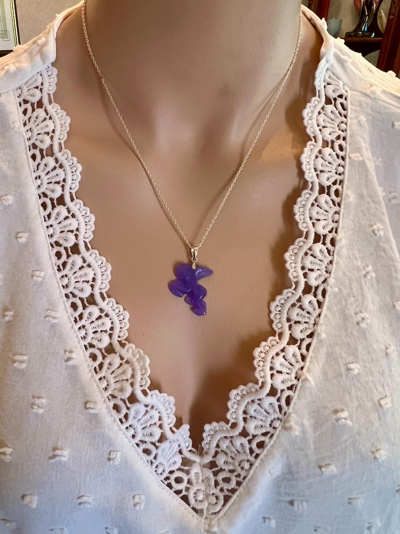 Puede incluir: Un collar de plata con un peque&ntilde;o colgante morado de forma abstracta. El colgante cuelga de una fina cadena de plata. El collar se lleva sobre una blusa blanca con un ribete de encaje detallado.