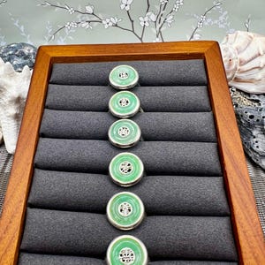 May include: A display of six jade rings with a silver band. Each ring features a circular, bright green jade stone with a silver filigree design in the center. The rings are arranged on a gray velvet display in a wooden box.