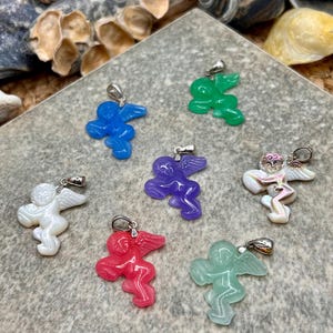 Puede incluir: Una colecci&oacute;n de colgantes de &aacute;ngeles coloridos con aros plateados. Los colgantes son de varios colores, incluyendo azul, verde, morado, rojo, blanco e iridiscente. Los &aacute;ngeles tienen alas y se exhiben sobre una superficie gris texturizada.