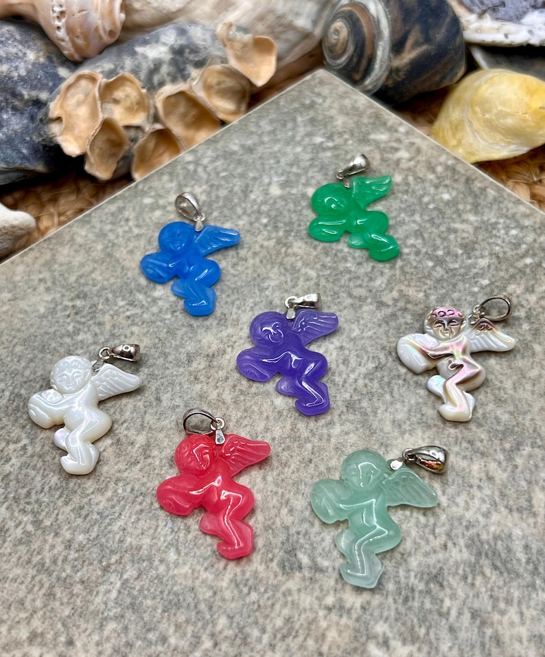 Puede incluir: Una colecci&oacute;n de colgantes de &aacute;ngeles coloridos con aros plateados. Los colgantes son de varios colores, incluyendo azul, verde, morado, rojo, blanco e iridiscente. Los &aacute;ngeles tienen alas y se exhiben sobre una superficie gris texturizada.