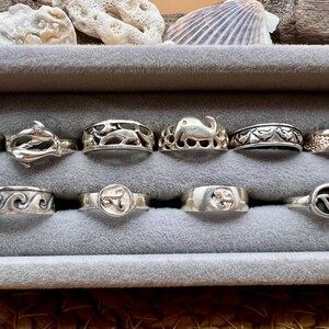 May include: A collection of silver rings with various designs, including waves, dolphins, and an elephant. The rings are displayed in a gray velvet jewelry box, with seashells in the background.