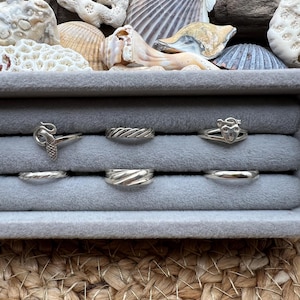 May include: A collection of silver rings displayed in a gray jewelry box. The rings feature various designs, including a mermaid, twisted bands, and heart-shaped motifs. Seashells are arranged in the background, adding a coastal theme.