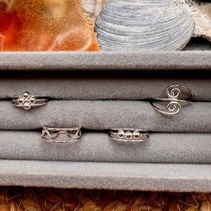 May include: A collection of silver rings displayed in a gray jewelry box. The rings feature various designs, including beaded clusters, wave patterns, and spiral swirls. The background includes a seashell and a piece of orange and white stone.