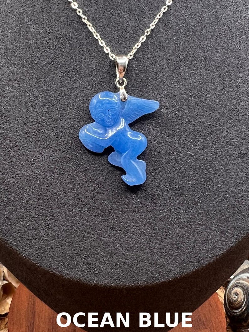 Puede incluir: Collar con colgante azul oc&eacute;ano con un &aacute;ngel tallado. El &aacute;ngel tiene alas extendidas y est&aacute; suspendido de una delicada cadena plateada. El colgante es de color azul vibrante. El texto "OCEAN BLUE" est&aacute; en la parte inferior.